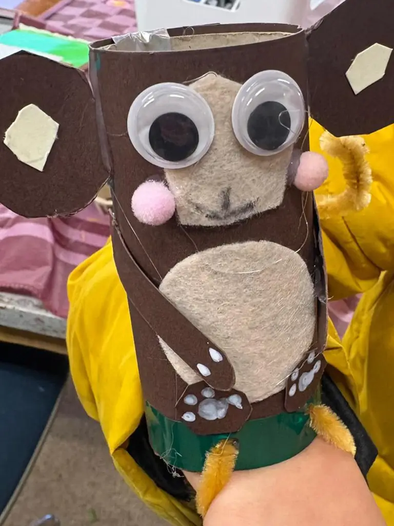 Een kind houdt een zelfgemaakte handpop van bruin karton en vilt met grote wiebeloogjes omhoog.