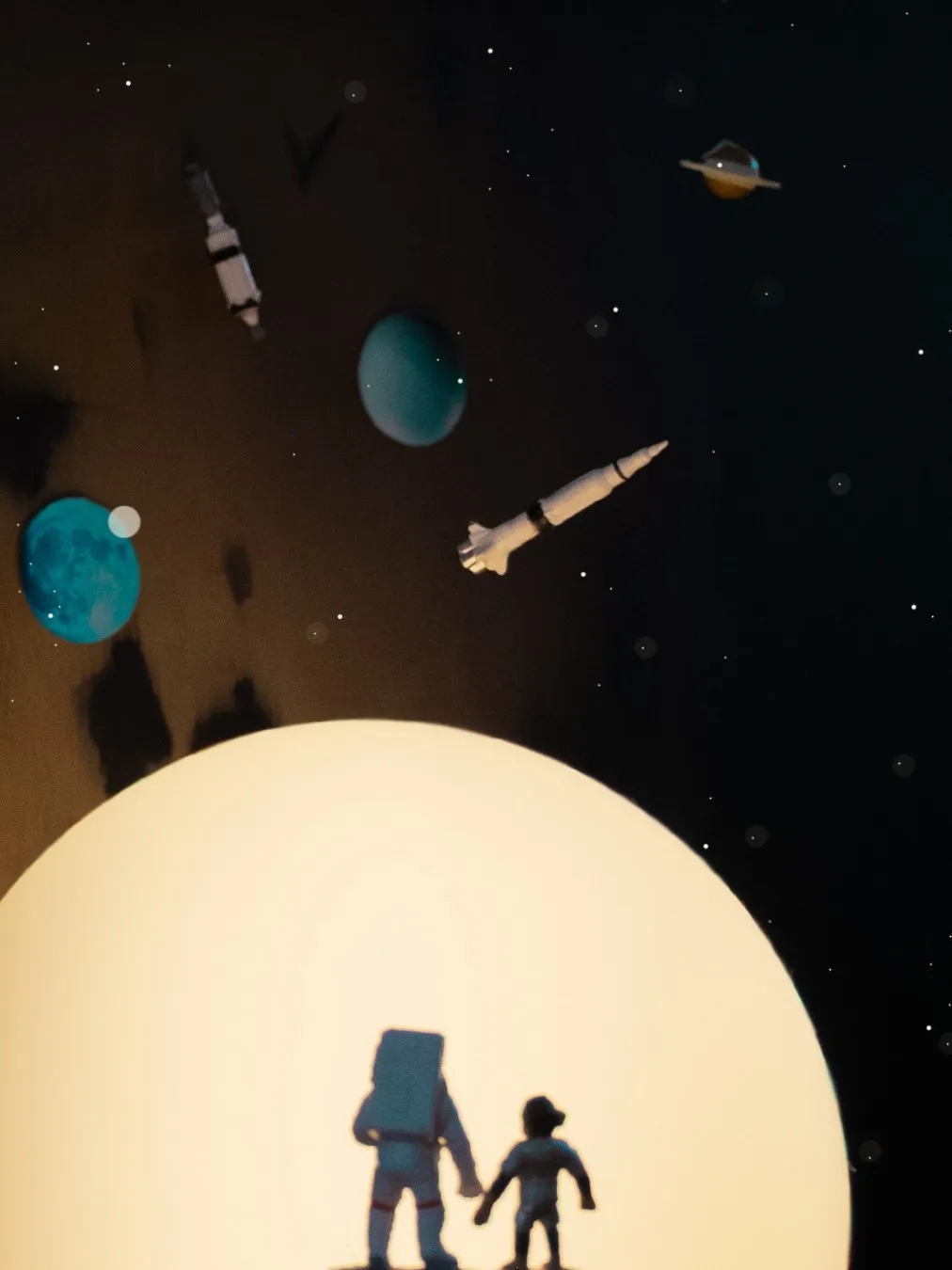 Een miniatuurscène van het heelal: twee astronautenfiguurtjes hand in hand op een lichtgevende planeet, met raketten, manen en Saturnus erboven