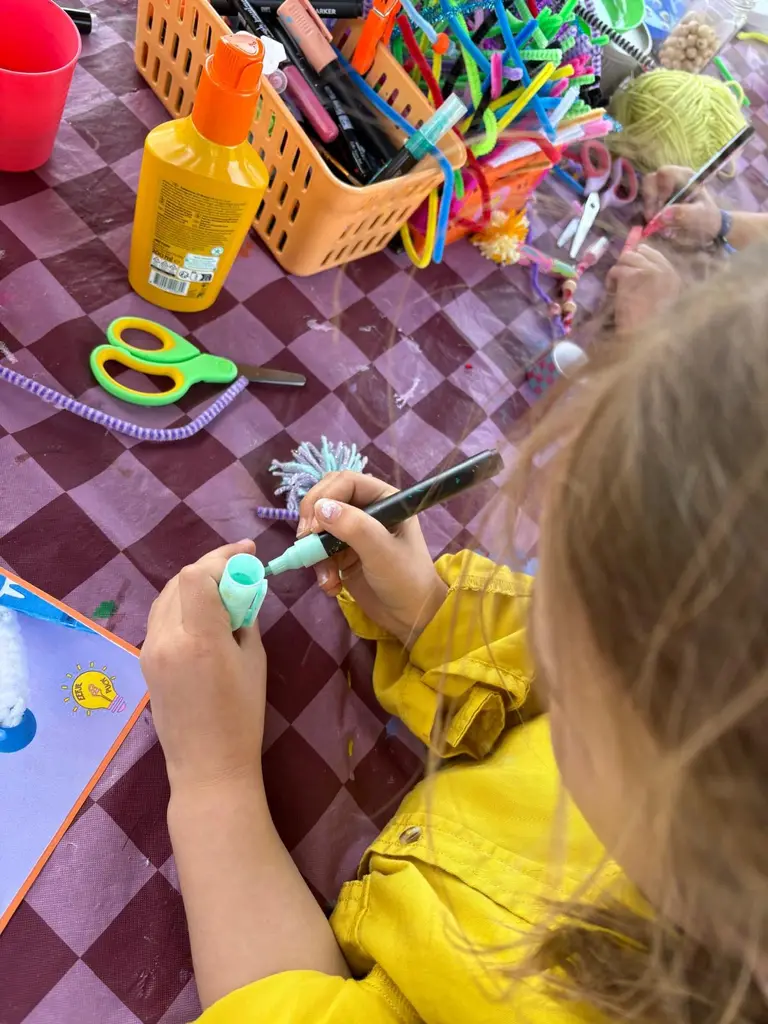 Een meisje in een gele jas beschildert een blauw pluizig figuurtje aan de knutseltafel.