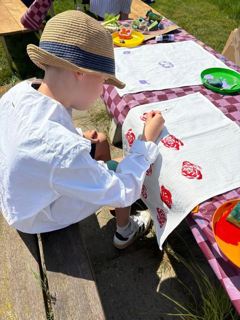 Een jongen met strohoed drukt figuren op een lap stof aan een picknicktafel buiten.
