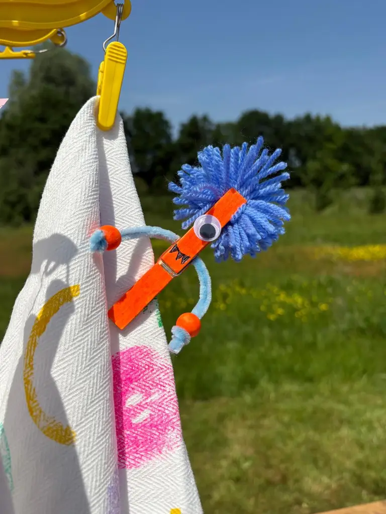 Een gekleurd vogeltje van pijpenragers en kralen hangt buiten aan een doek.
